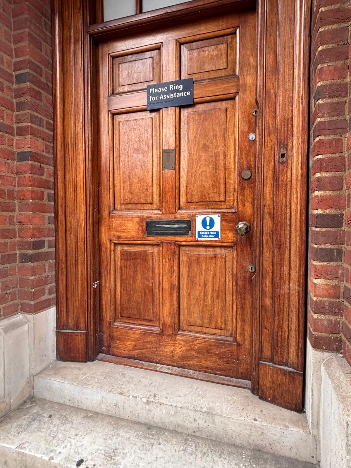 Heritage timber communal entrance door with compliant fire-safety signage — blue 'Escape route, keep clear' notice and brass 'Please ring for assistance' plate.